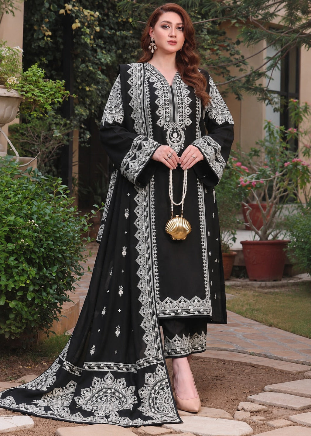 A woman stands outdoors on a stone path wearing a long black embroidered dress with intricate silver-white floral and geometric patterns. She holds a small gold shell-shaped handbag and wears beige heels, with green plants and a house visible in the background.