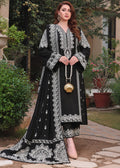 A woman stands outdoors on a stone path wearing a long black embroidered dress with intricate silver-white floral and geometric patterns. She holds a small gold shell-shaped handbag and wears beige heels, with green plants and a house visible in the background.