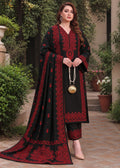 A woman stands outdoors wearing a black traditional outfit with rich red embroidery, holding a matching embroidered dupatta. The setting features a stone pathway and lush green plants.