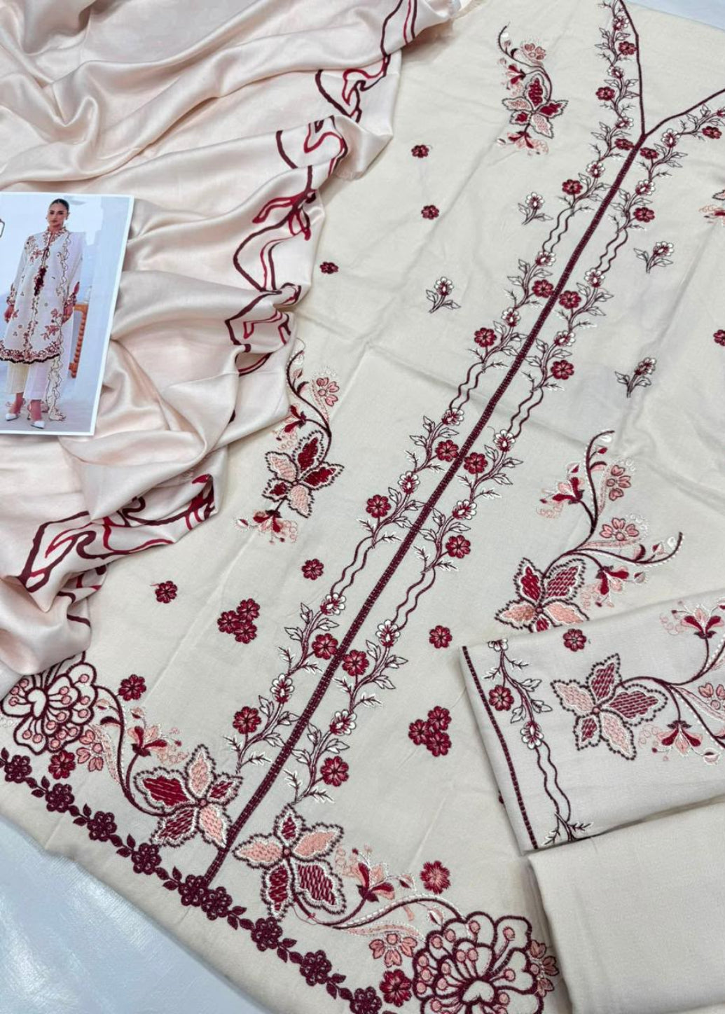 Cream-colored traditional embroidered fabric with pink and maroon floral patterns, draped alongside a matching satin dupatta and a small printed outfit card.
