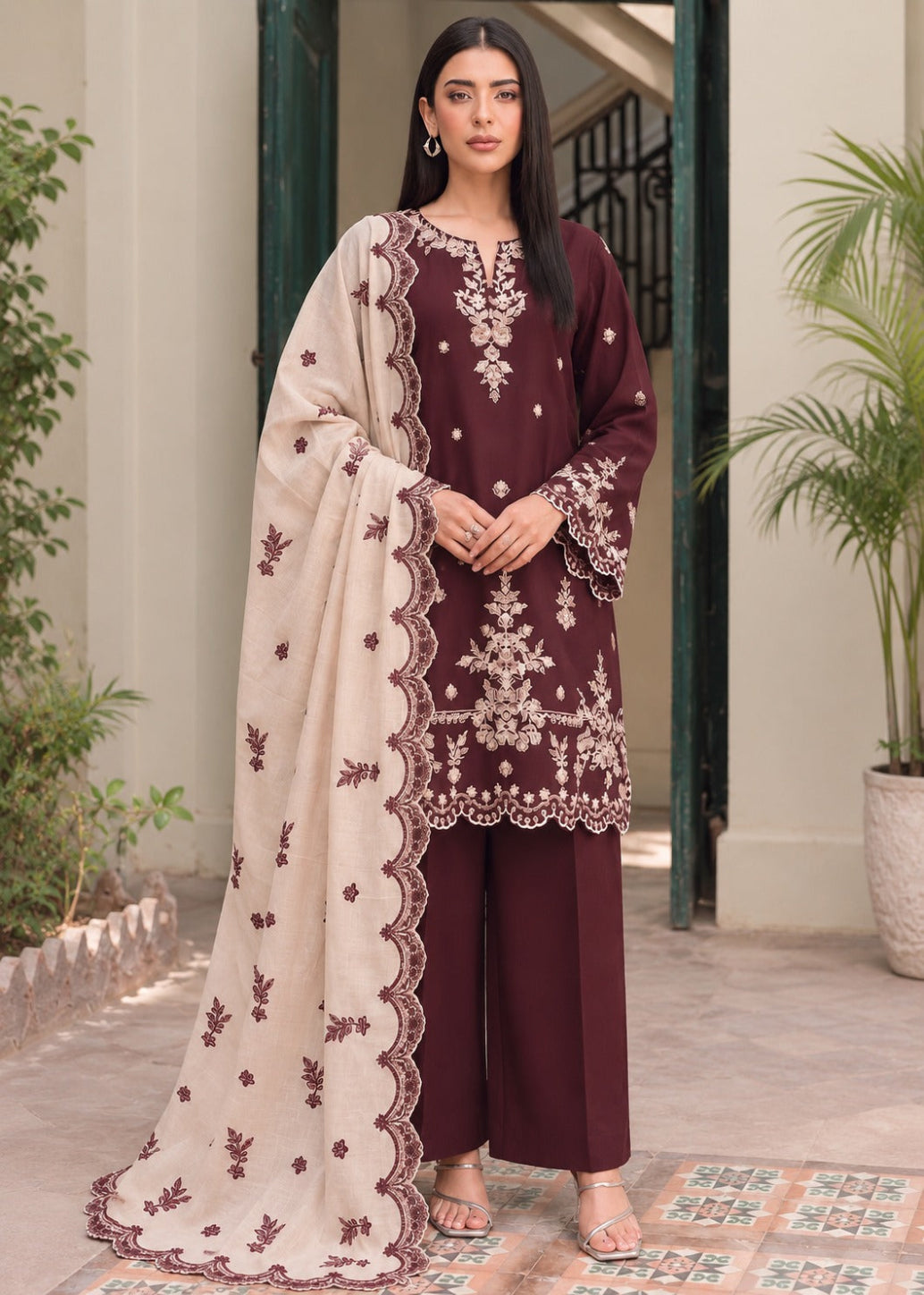 A woman models a three-piece traditional South Asian salwar kameez suit. The kurta is deep maroon with intricate cream and gold embroidery, paired with matching maroon wide-leg trousers and a cream dupatta featuring a scalloped maroon border and embroidered motifs.