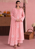 A woman stands in a pink room wearing a long light pink traditional outfit with white printed motifs and matching pants. The setting includes a pink wall, a table with bottles, a wicker basket, and flowers.