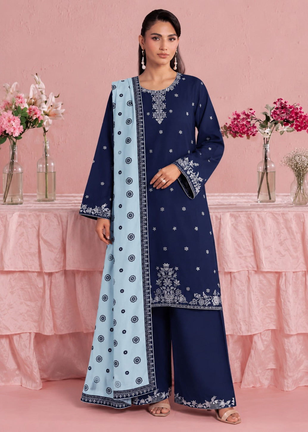 A woman models a navy blue traditional Indian salwar kameez suit with a light blue dupatta, set against a pink backdrop with decorative floral arrangements.