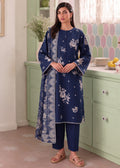 A woman modeling a navy blue three-piece Indian ethnic suit with white floral and paisley prints, standing in a pastel green kitchen setting.