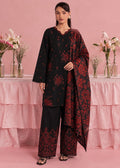 A woman models a black three-piece traditional outfit with red floral embroidery, featuring a long kurta, wide-leg trousers, and a matching dupatta, set against a pink backdrop with decorative floral arrangements.