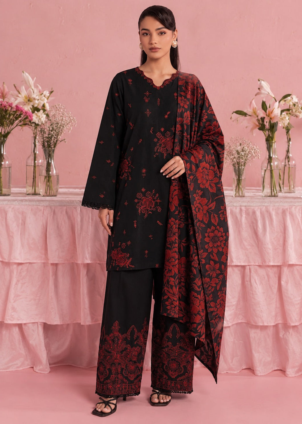 A woman models a black three-piece traditional outfit with red floral embroidery, featuring a long kurta, wide-leg trousers, and a matching dupatta, set against a pink backdrop with decorative floral arrangements.