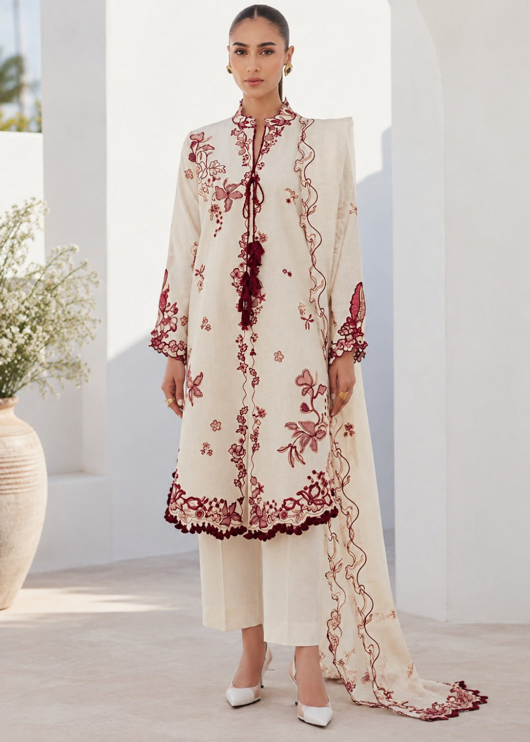 A woman models a traditional three-piece salwar kameez suit in cream with deep red floral embroidery and border detailing, set against a bright, sunlit white architectural background.