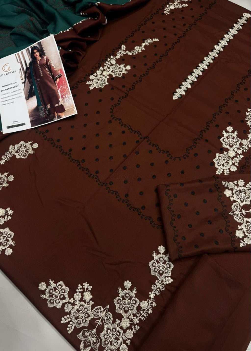 A brown ethnic salwar suit fabric set with intricate white floral embroidery and black polka dots, shown with a matching folded dupatta and a brand card.