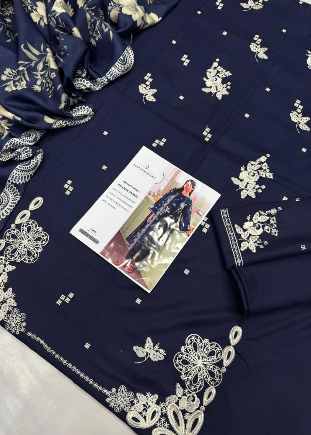 A navy blue fabric garment or textile laid flat, decorated with white floral and geometric embroidery. A folded matching fabric piece with embroidered border is placed on top, along with a printed card and a small matching accessory.