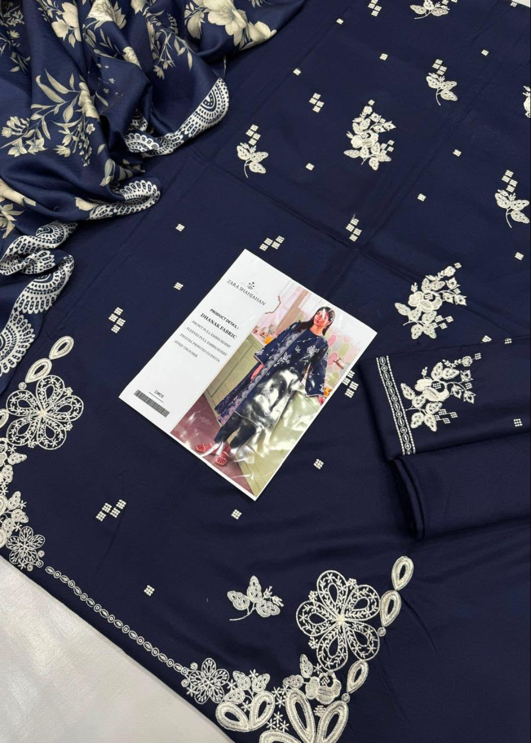 A navy blue fabric garment or textile laid flat, decorated with white floral and geometric embroidery. A folded matching fabric piece with embroidered border is placed on top, along with a printed card and a small matching accessory.