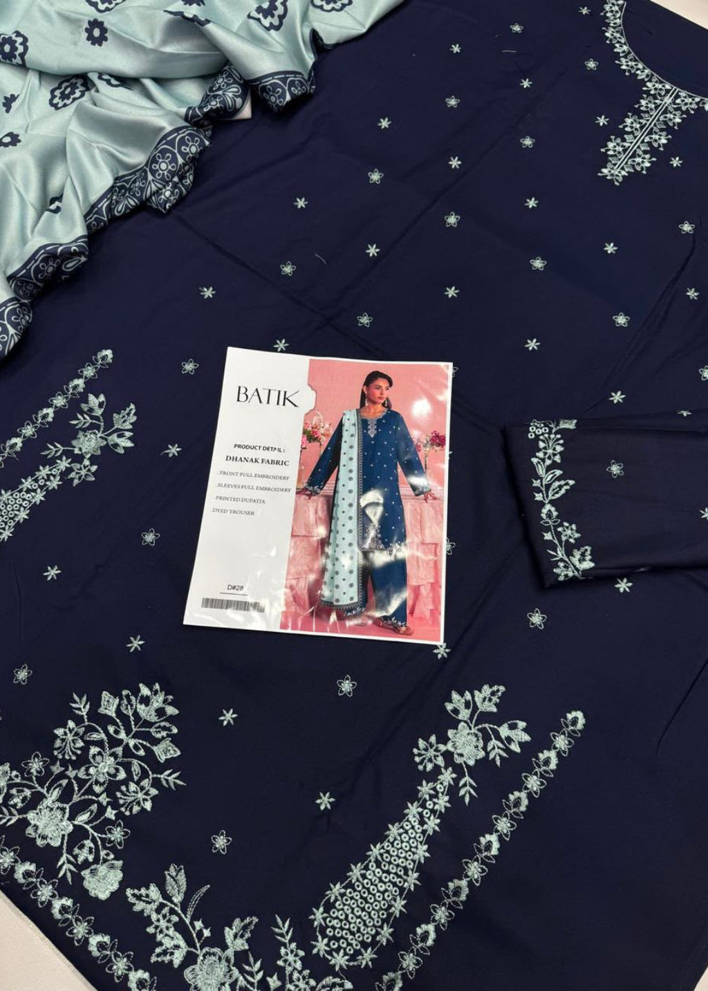 A navy blue embroidered salwar kameez suit fabric set with a matching light blue dupatta, featuring white floral embroidery and small star motifs. A BATIK brand card is placed on top of the fabric.