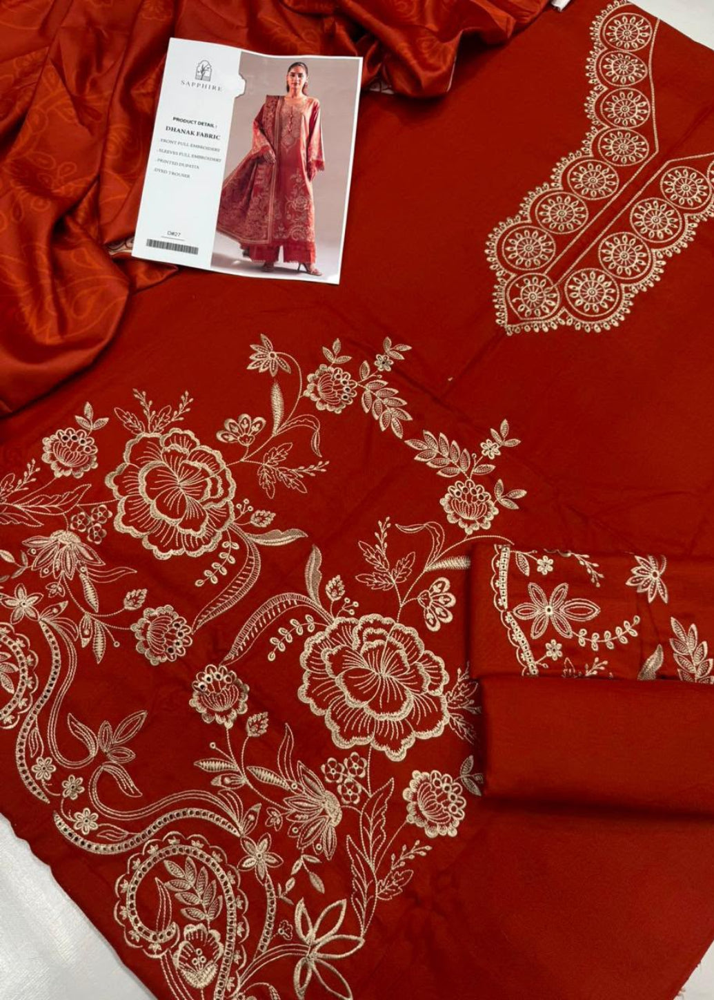A deep red or maroon unstitched salwar suit fabric set with intricate gold floral embroidery, shown alongside a matching silk-like dupatta and a printed garment card.