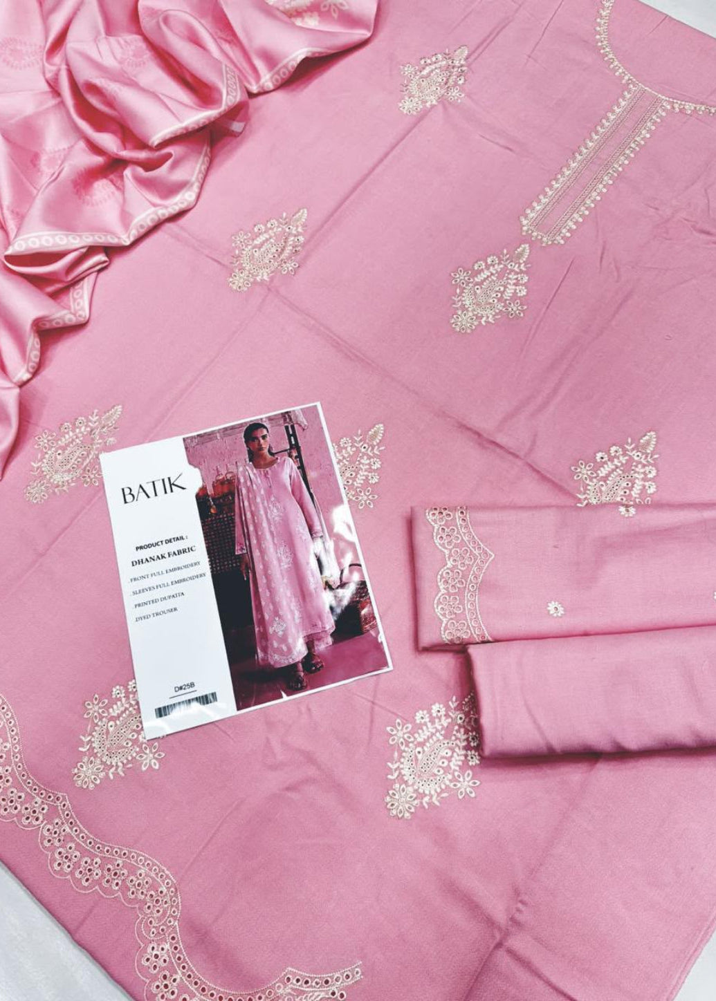 Pink fabric clothing set laid flat with embroidered motifs, a matching satin-like draped piece, folded matching fabric, and a printed card featuring a model wearing the garment.