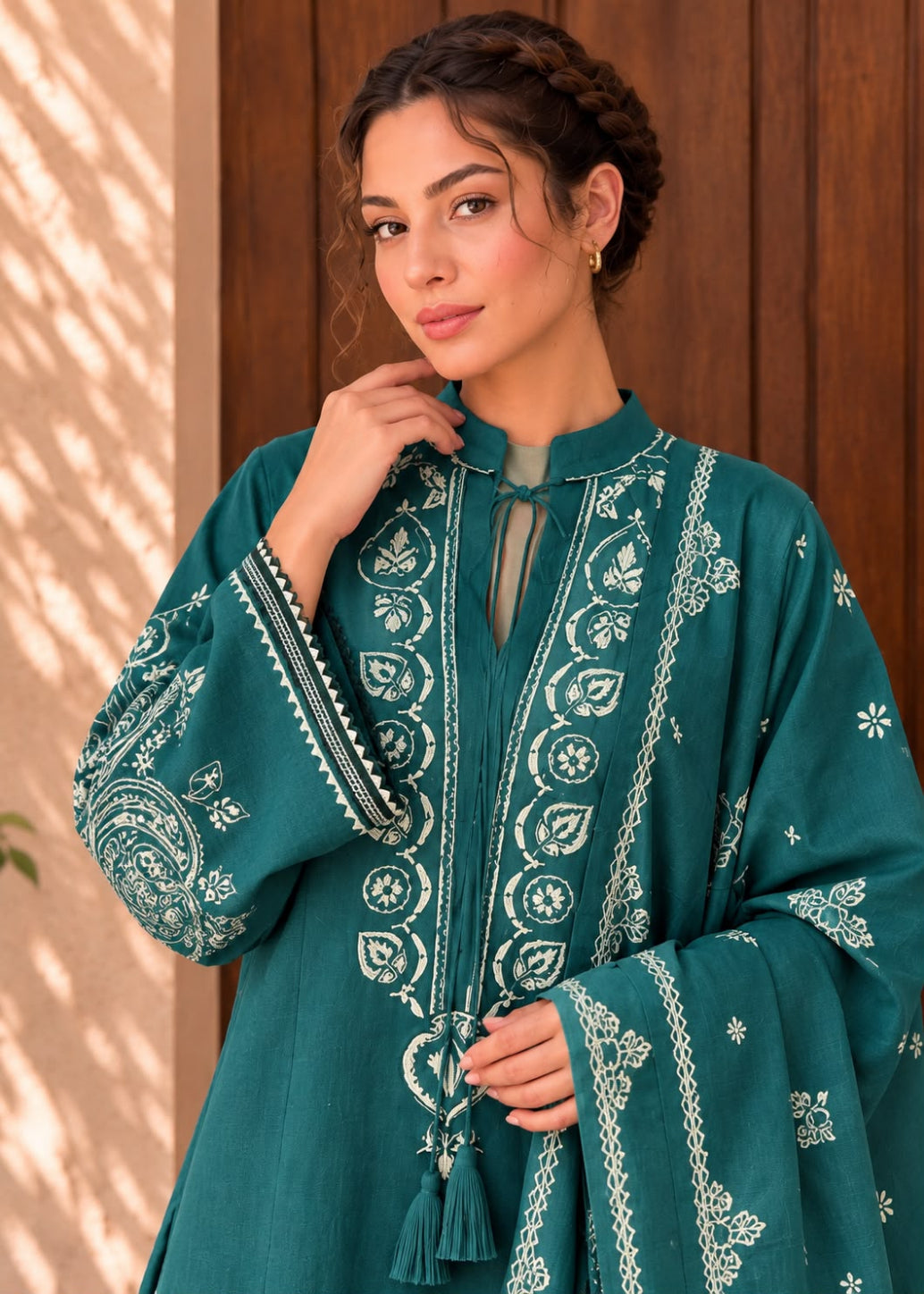 A woman wearing a teal green embroidered traditional kurta with white floral and geometric patterns, standing against a wooden door background.