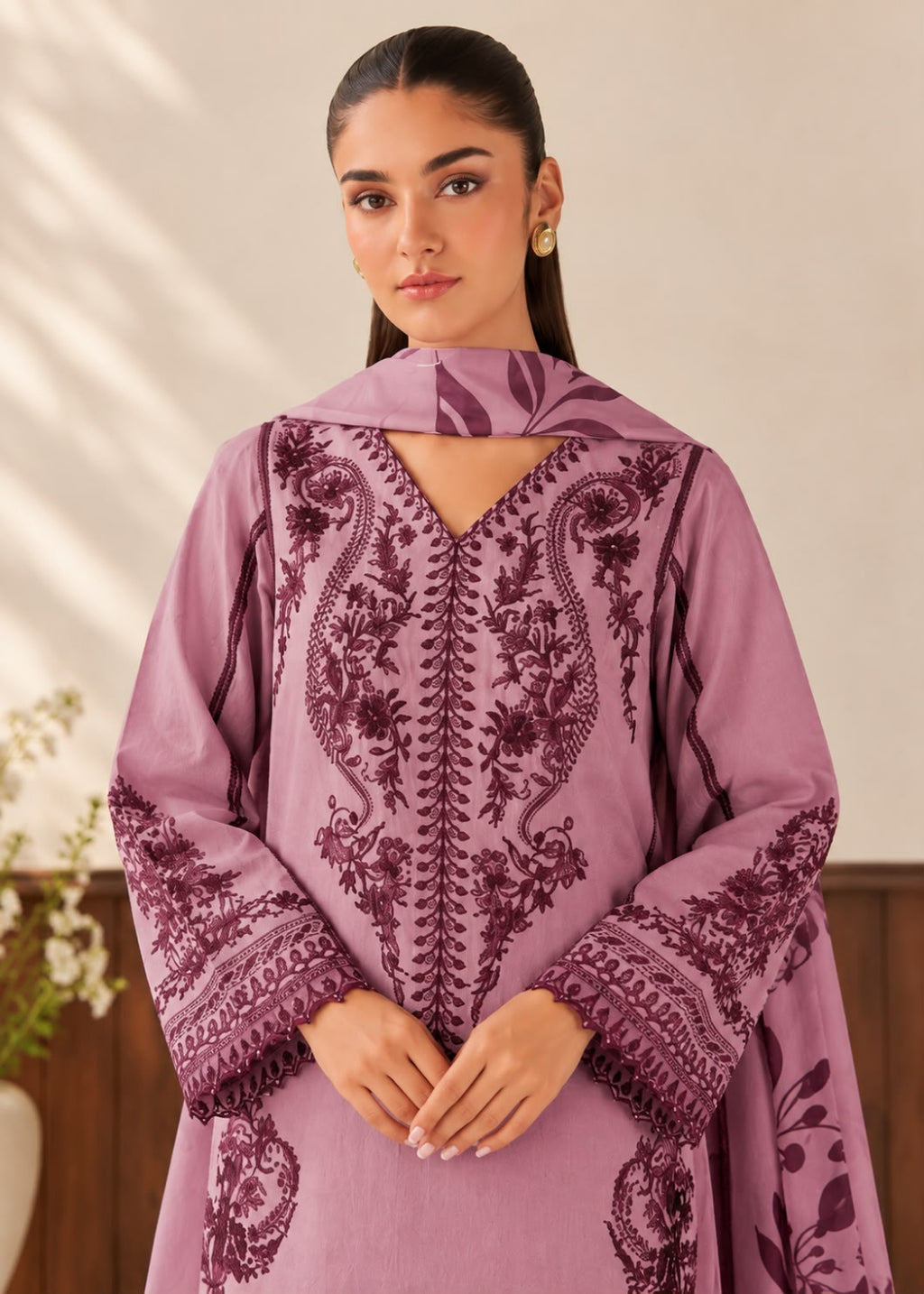 A woman wearing a lavender purple embroidered tunic with dark purple floral and paisley embroidery, standing against a warm beige background.
