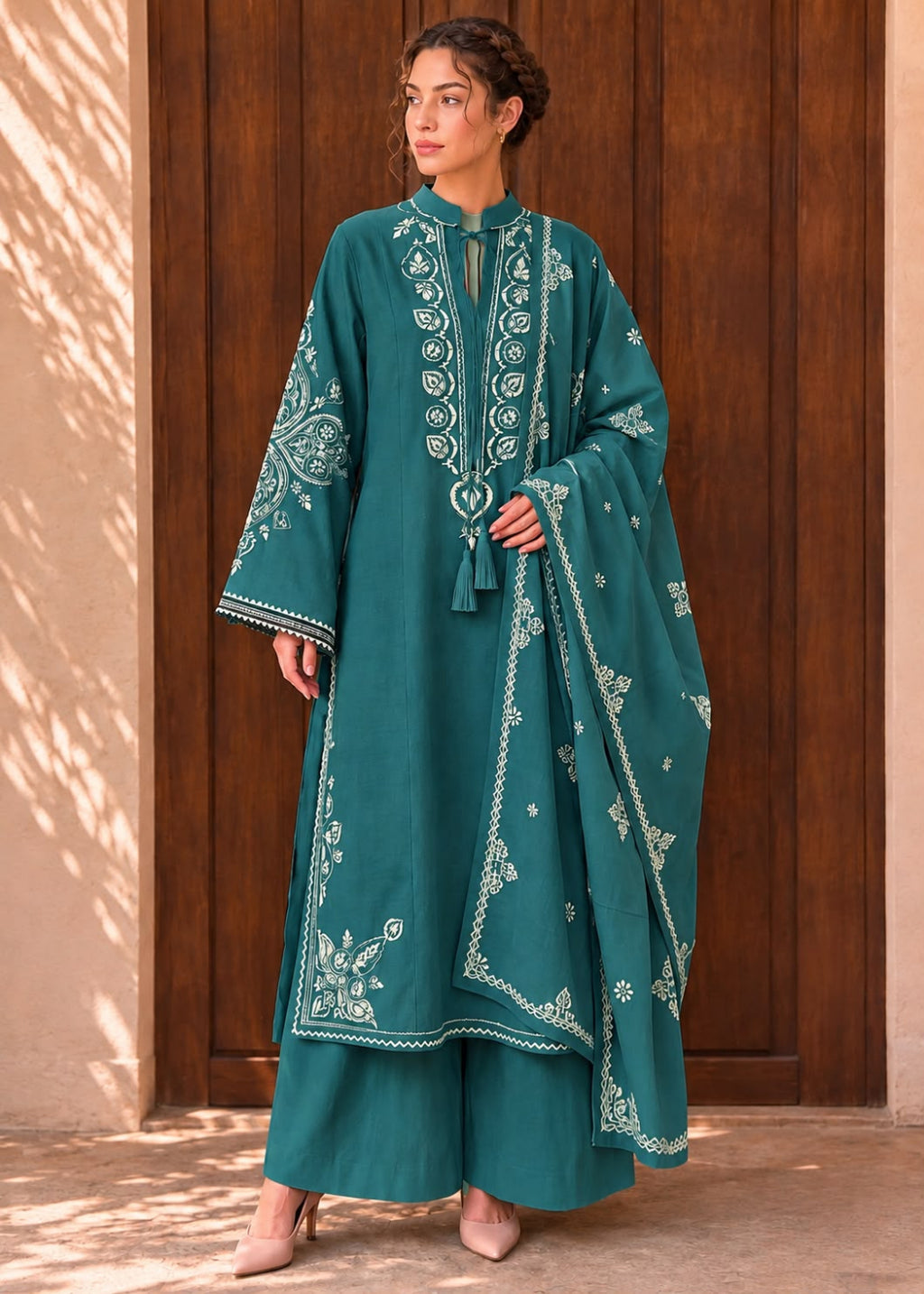 A woman models a teal green traditional embroidered kurta set with matching wide-leg trousers and a dupatta, standing in front of a dark wooden door.