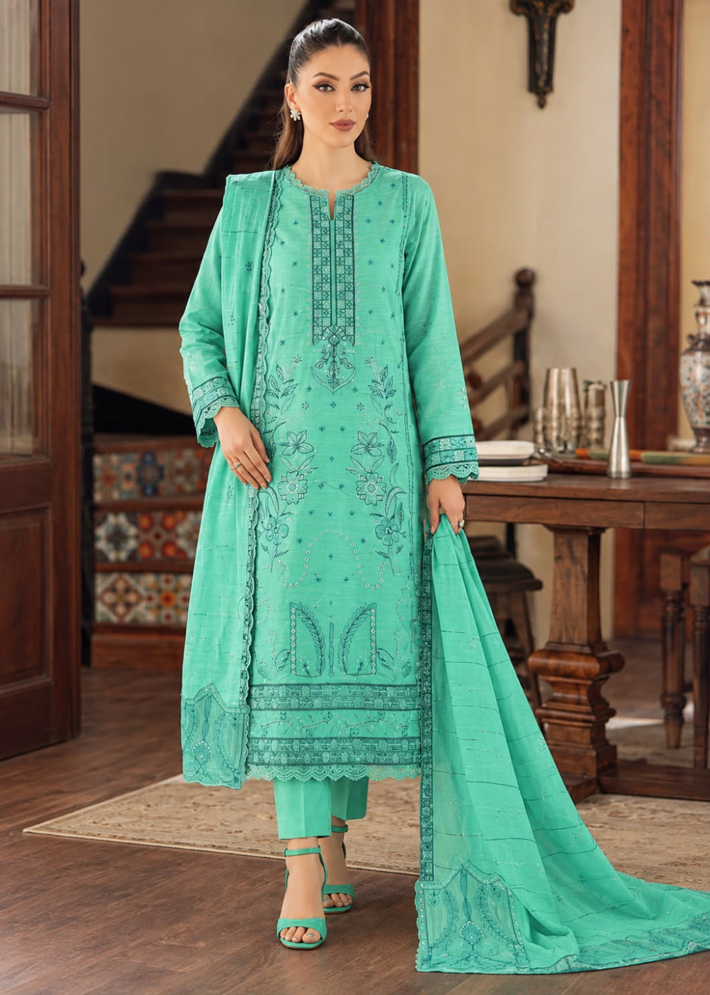 A woman models a mint green traditional South Asian salwar kameez suit with matching dupatta in an indoor wooden staircase setting. The suit features detailed floral embroidery and prints on the front, matching pants, and a long dupatta.