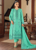 A woman models a mint green traditional South Asian salwar kameez suit with matching dupatta in an indoor wooden staircase setting. The suit features detailed floral embroidery and prints on the front, matching pants, and a long dupatta.