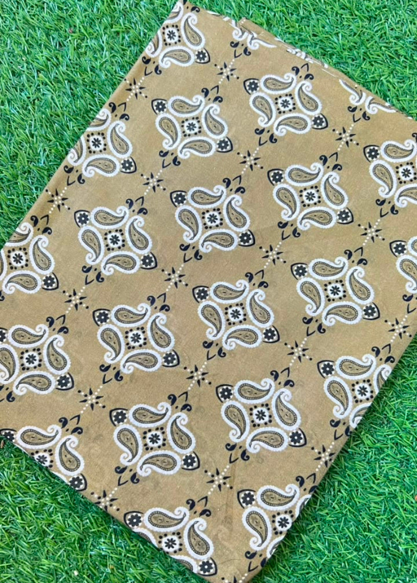 A rectangular fabric item, likely a bandana or scarf, featuring a brown, white, and black paisley pattern, laid flat on a green grass background.