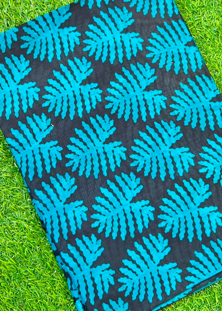 A close-up of a black fabric with a bright turquoise blue leaf or fern-like block print pattern, laid flat on green grass.