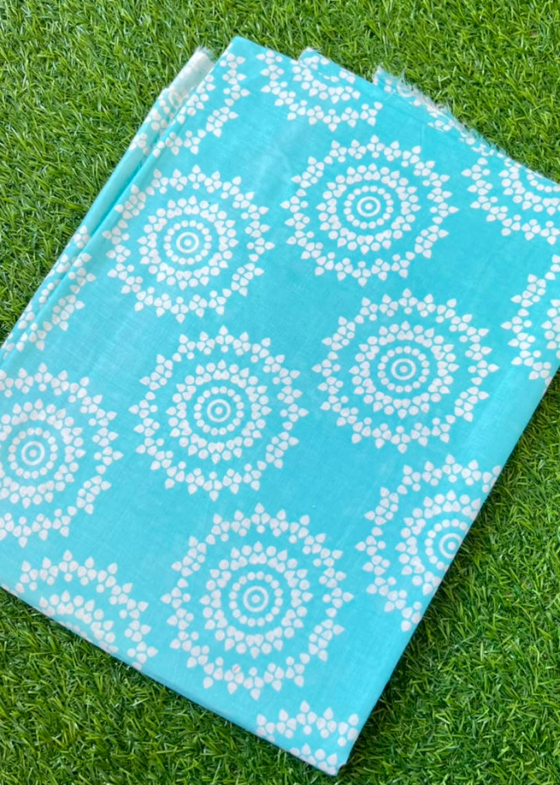 A folded piece of turquoise blue fabric with a white mandala pattern, laid flat on green grass.