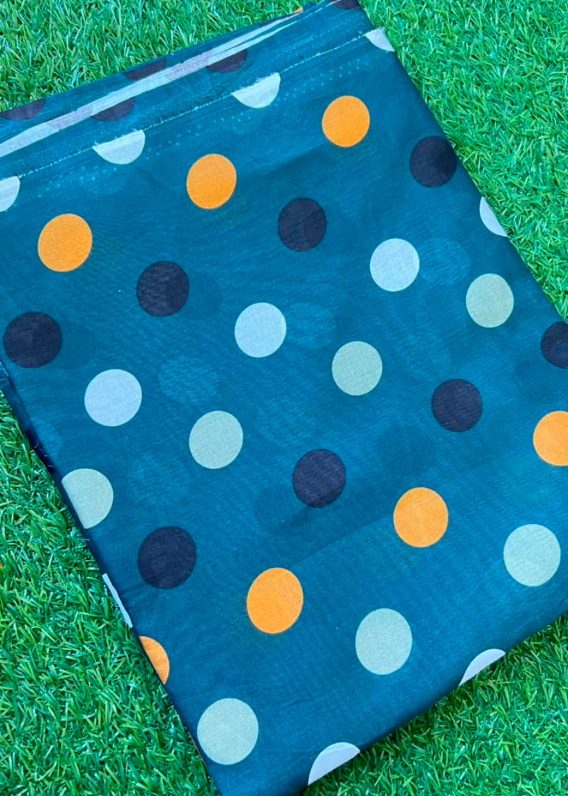 A folded dark teal blue fabric item, likely a scarf or shawl, featuring a pattern of large polka dots in orange, black, light blue, and gray, displayed on a green grass background.