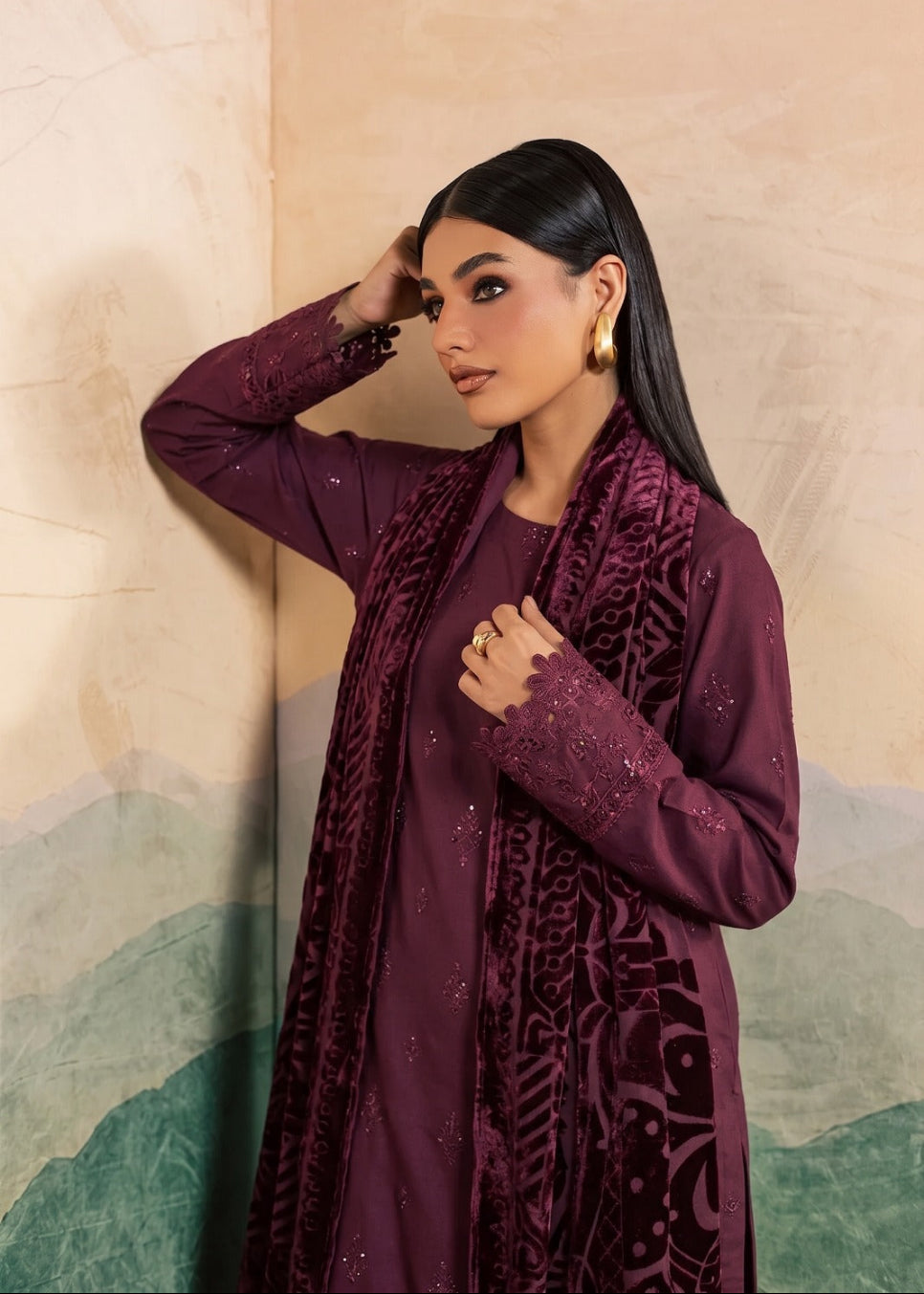 A woman modeling a deep plum purple traditional outfit with matching embroidered scarf, standing against a beige and green mountain-patterned wall.