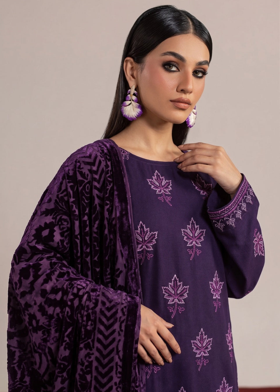 A woman modeling a deep purple traditional outfit with matching embroidered floral motifs and a patterned draped shawl, set against a plain beige studio background.