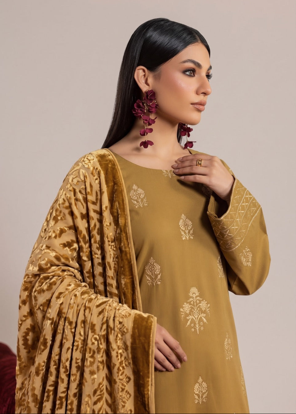 A woman modeling a traditional Indian salwar kameez suit in mustard yellow, featuring a kurta with delicate gold floral embroidery, a matching embroidered dupatta, and large maroon floral earrings, set against a solid beige background.