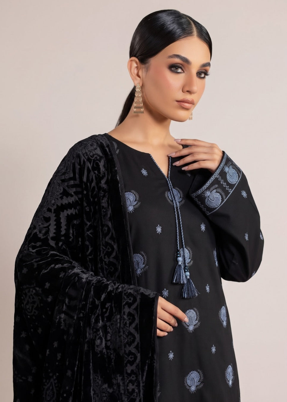 A woman modeling a black embroidered kurta with a matching black velvet shawl draped over her shoulders, set against a solid light beige background.
