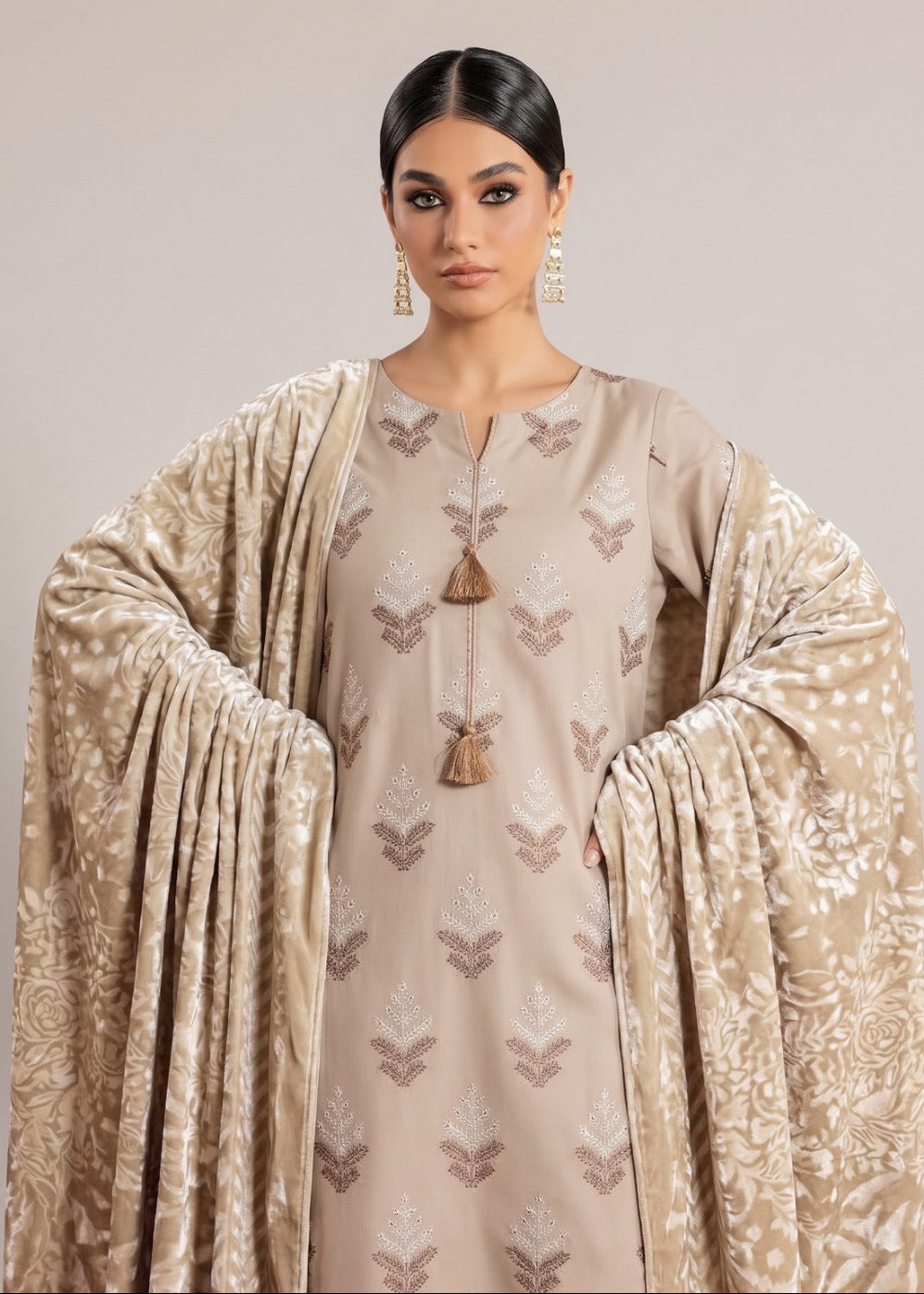 A woman modeling a beige traditional outfit with a matching patterned dupatta, featuring embroidered motifs and a tassel detail at the neckline, set against a solid light beige background.
