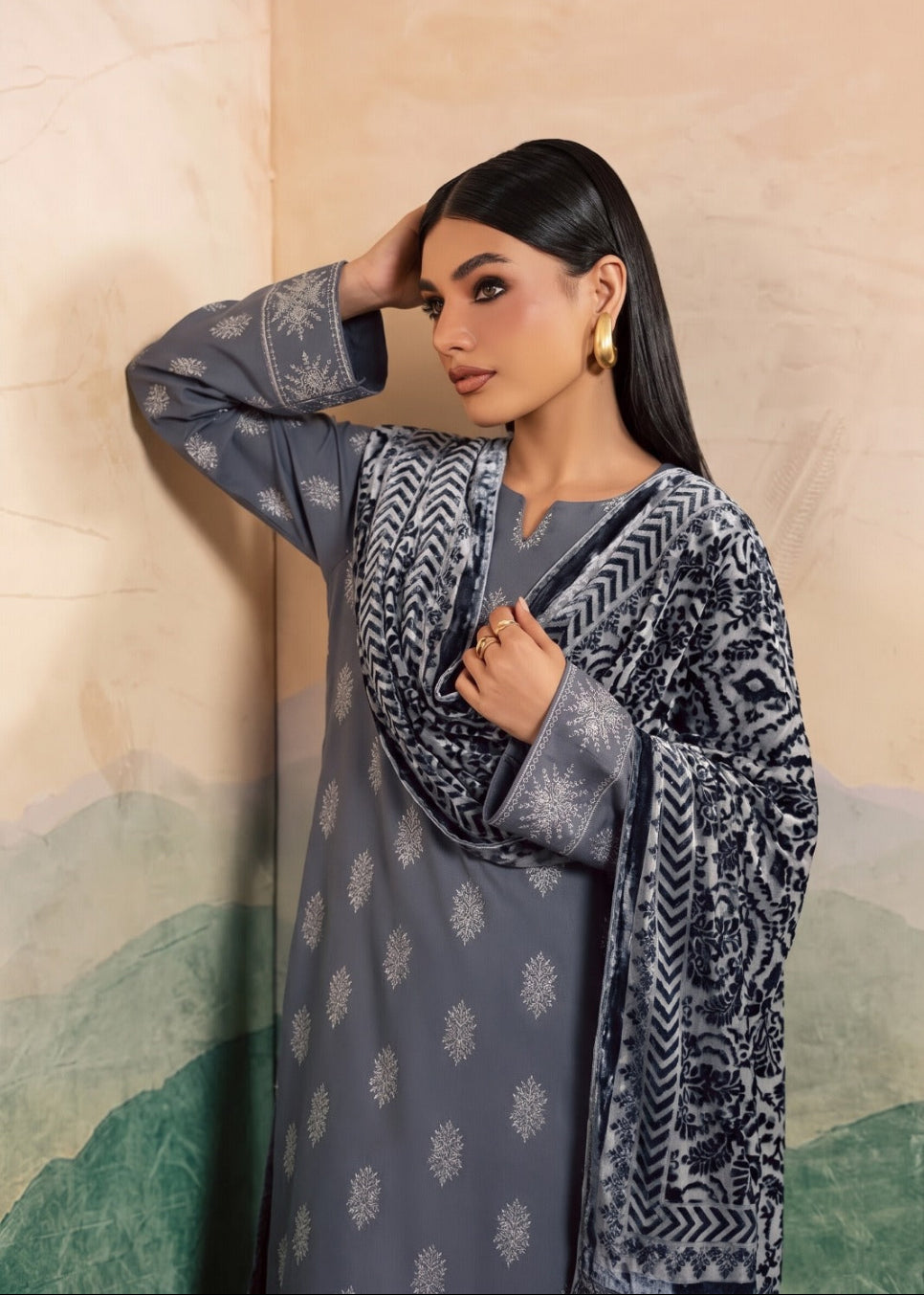 A woman modeling a three-piece traditional outfit featuring a slate gray kurta with white motifs, a matching patterned inner layer, and a draped dupatta with geometric patterns, set against a beige and green scenic backdrop.