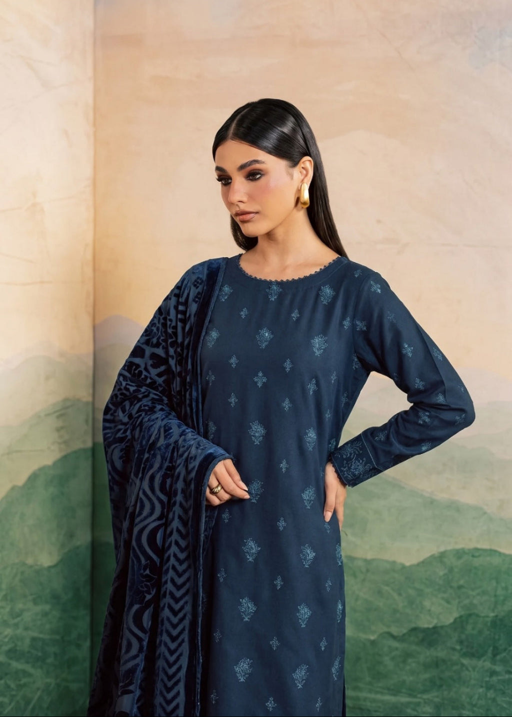 A woman modeling a dark navy blue long-sleeve kurta with small light blue floral prints, paired with a matching navy dupatta featuring a geometric pattern, set against a scenic backdrop of mountains and a warm-toned wall.