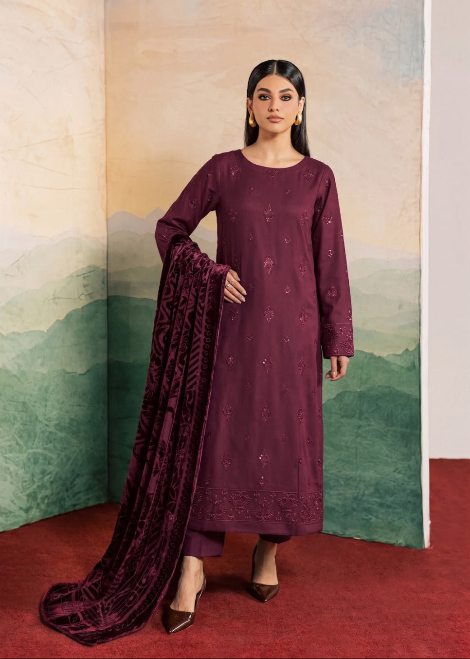 A woman models a deep plum purple traditional Indian salwar kameez suit with matching embroidered kurta, trousers, and a long dupatta, set against a scenic painted backdrop.