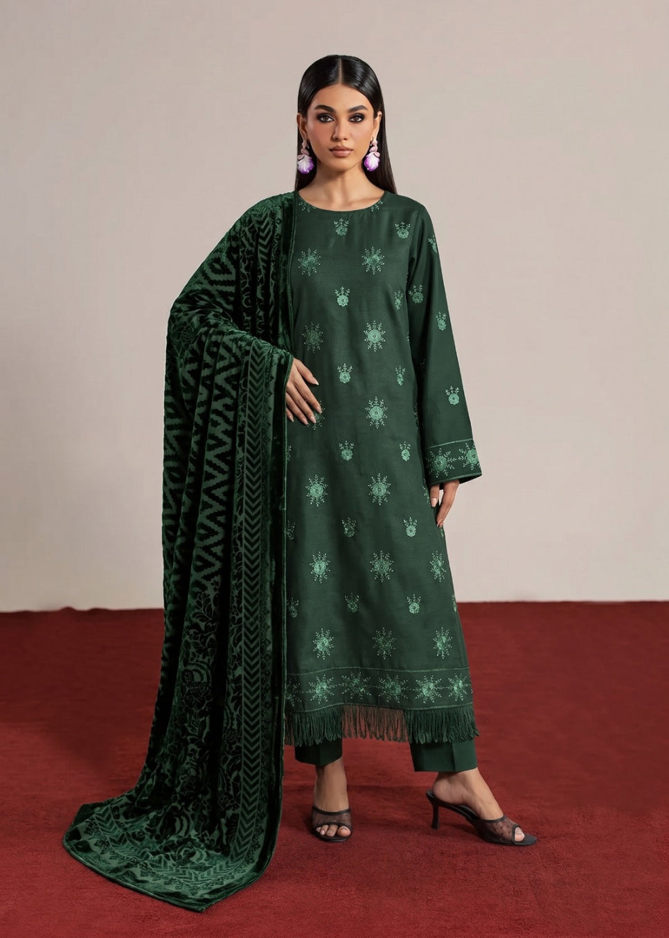 A woman models a dark green traditional outfit with matching printed dupatta against a neutral beige wall and red carpet.