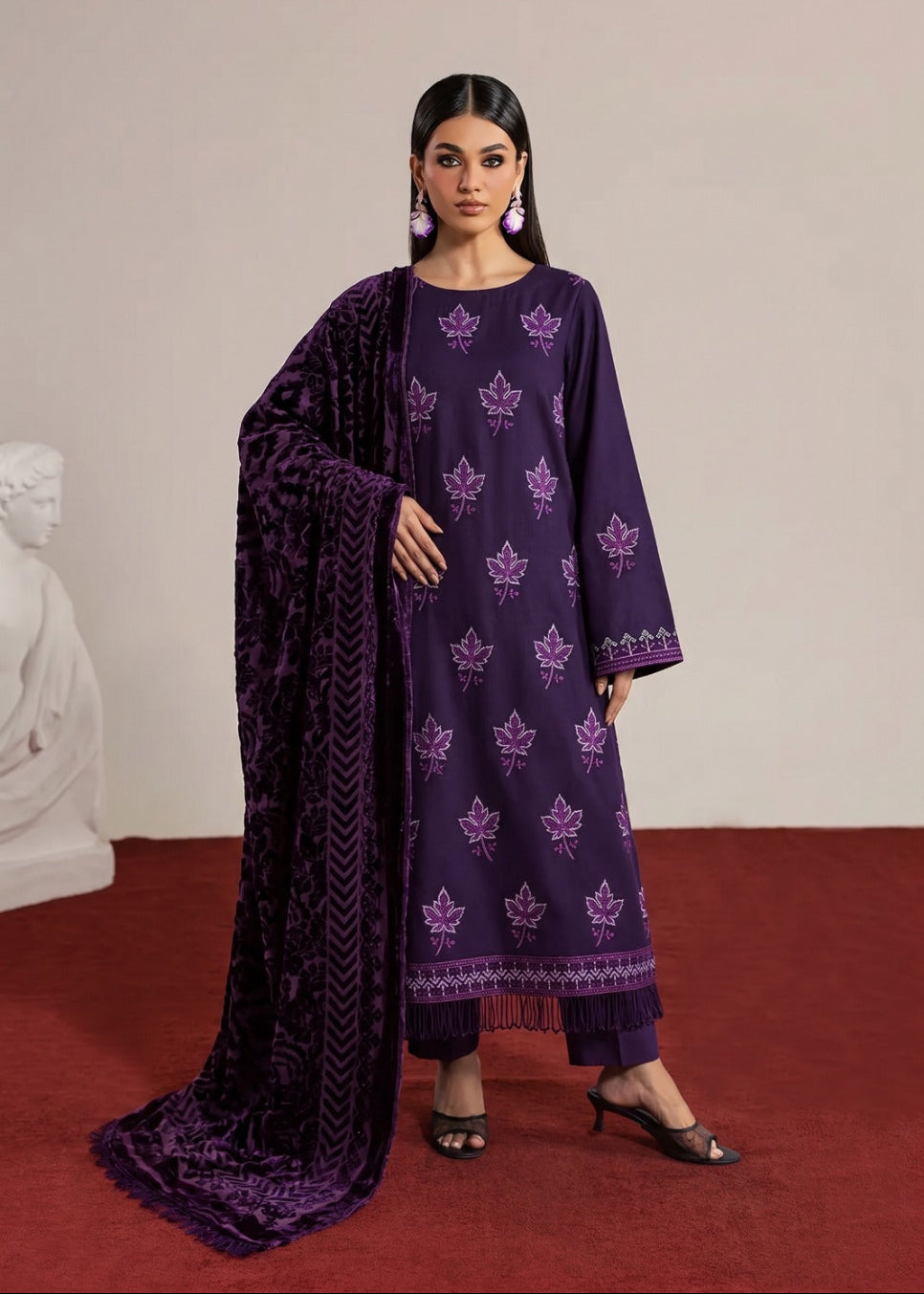 A woman models a deep purple three-piece traditional outfit with matching embroidered kurta, pants, and a textured dupatta, set against a minimalist beige wall and red carpet.