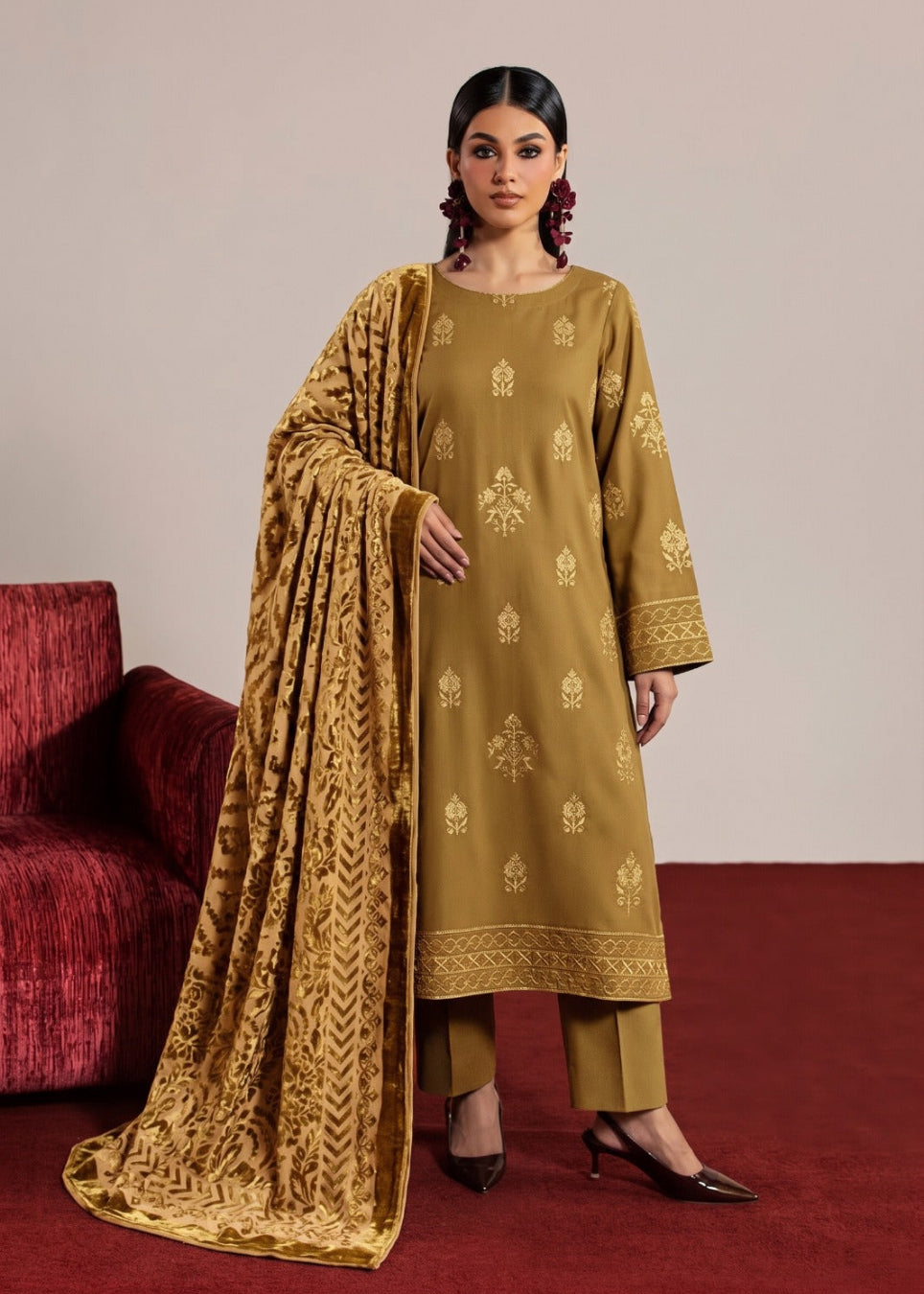 A woman models a traditional Indian salwar kameez suit in a warm mustard yellow color with gold zari embroidery, paired with matching trousers and a heavily embroidered dupatta, set against a neutral studio background with a red velvet chair.