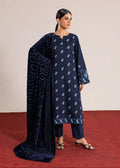 A woman modeling a navy blue traditional outfit with white floral embroidery, matching navy pants, and a patterned navy dupatta, standing on a red carpet against a beige studio backdrop.