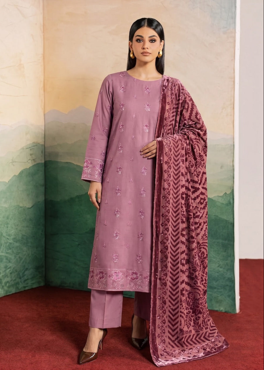 A woman models a traditional Indian outfit consisting of a dusty purple kurta with floral embroidery, matching trousers, and a patterned mauve dupatta, set against a scenic painted backdrop.