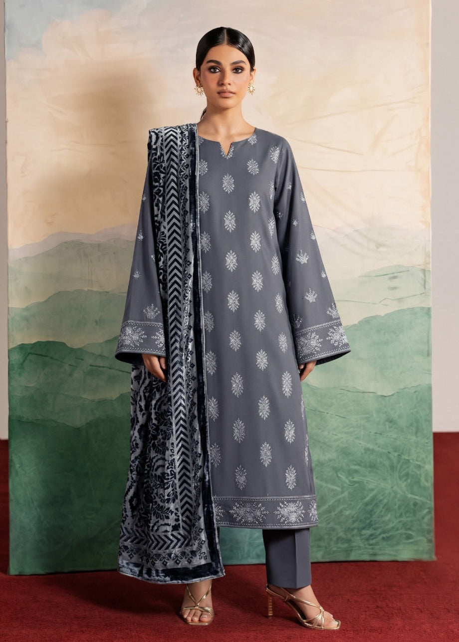A woman models a three-piece traditional Indian outfit consisting of a gray kurta with white motifs, matching gray trousers, and a navy blue patterned dupatta, set against a scenic painted backdrop.