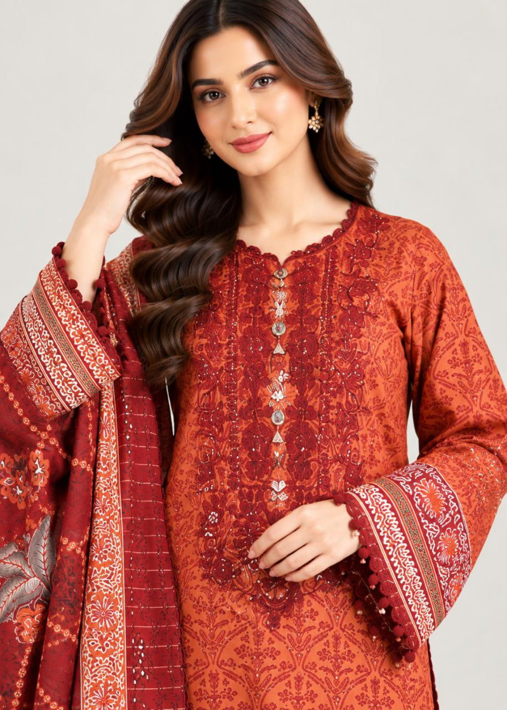 A woman modeling a rust-orange and deep red embroidered kurta with matching patterned dupatta against a light gray studio background.