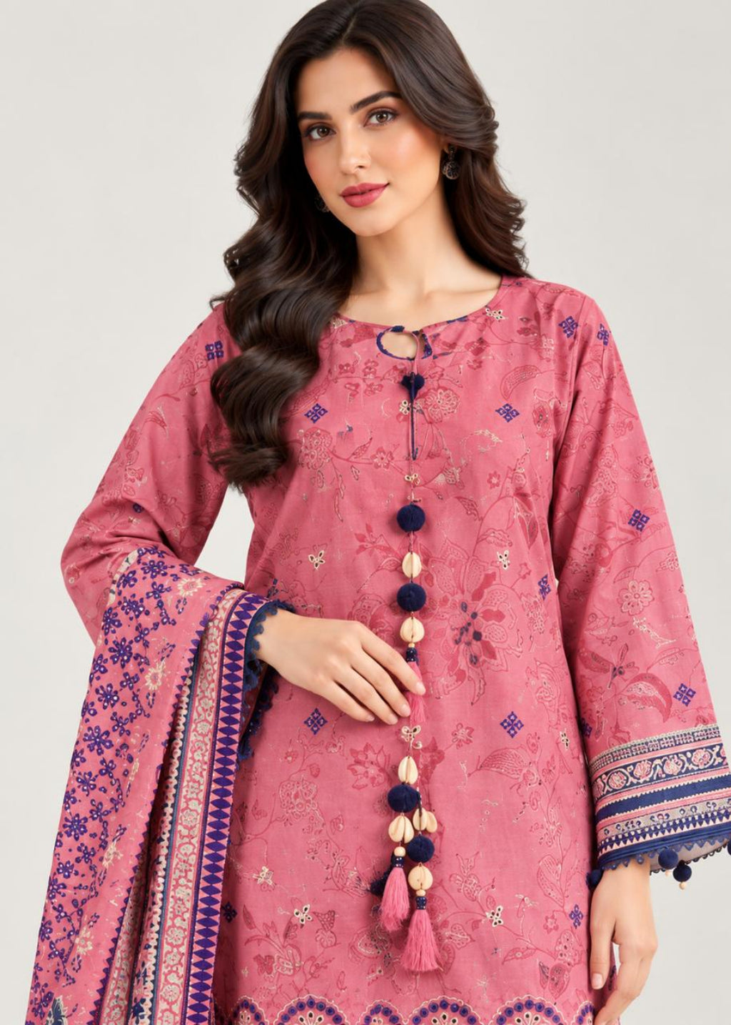 A woman modeling a pink embroidered kurta with a matching patterned dupatta, featuring navy and beige tassel details, set against a plain light gray background.