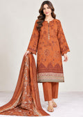 A woman models a rust-orange 3-piece salwar kameez suit set with matching printed dupatta. The kurta features dark brown floral patterns and a decorative border, paired with matching trousers and a dupatta with gold and brown embroidery.