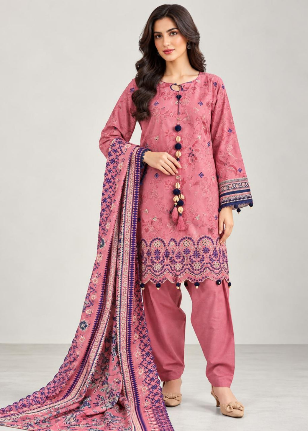 A woman models a pink traditional South Asian outfit with matching pants and a long patterned dupatta against a plain light gray studio background.