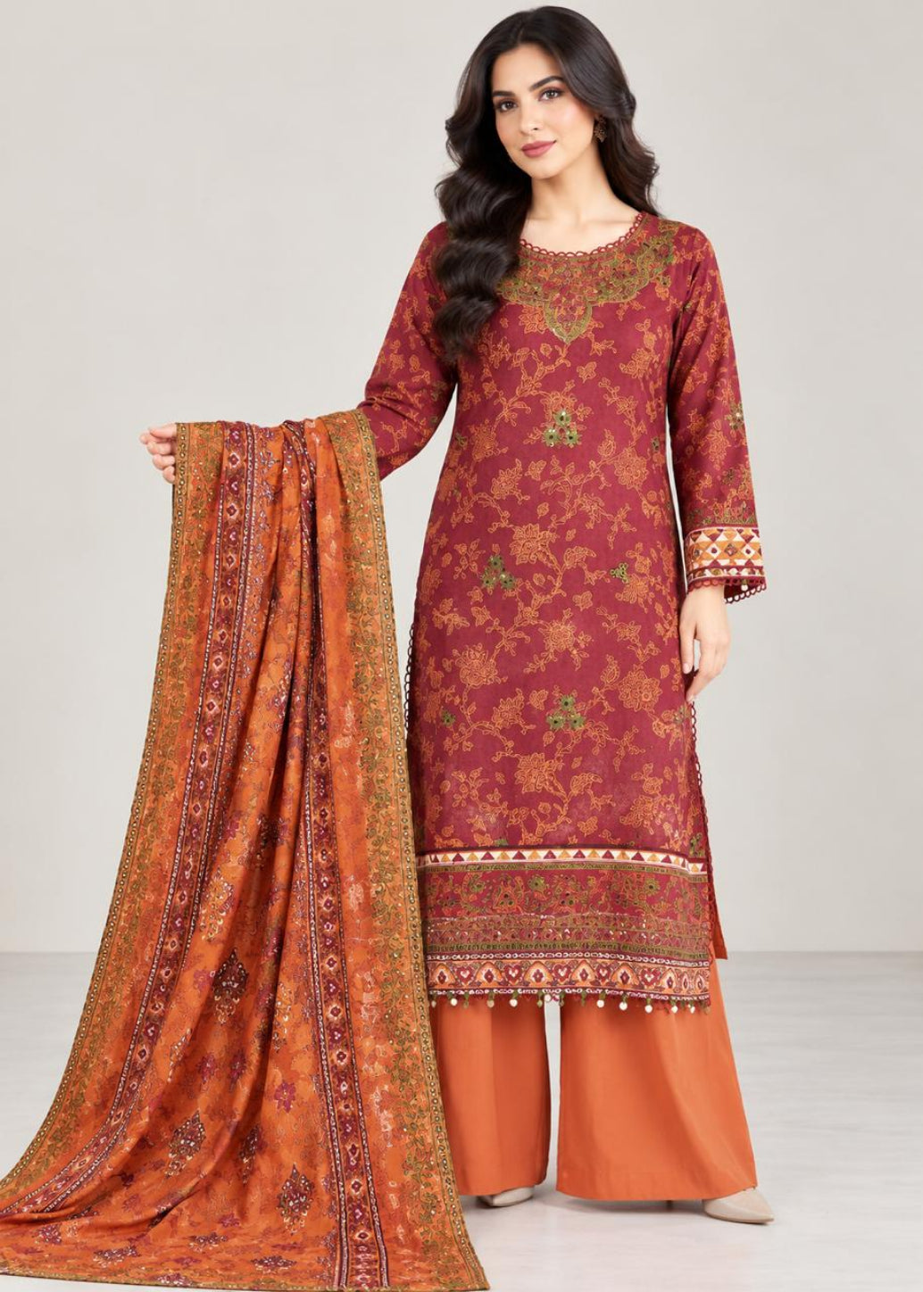 A woman models a traditional South Asian salwar kameez suit set. The set features a deep maroon kameez kurta with gold and green floral embroidery, paired with wide-leg rust-orange trousers and a matching rust-orange dupatta with intricate gold and green border patterns.