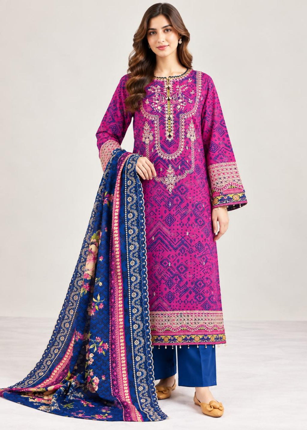 A woman modeling a traditional three-piece salwar kameez suit. The kurta is a vibrant magenta pink with intricate gold and silver embroidery and geometric patterns, paired with blue pants and a matching blue dupatta featuring a floral print and ornate gold border.