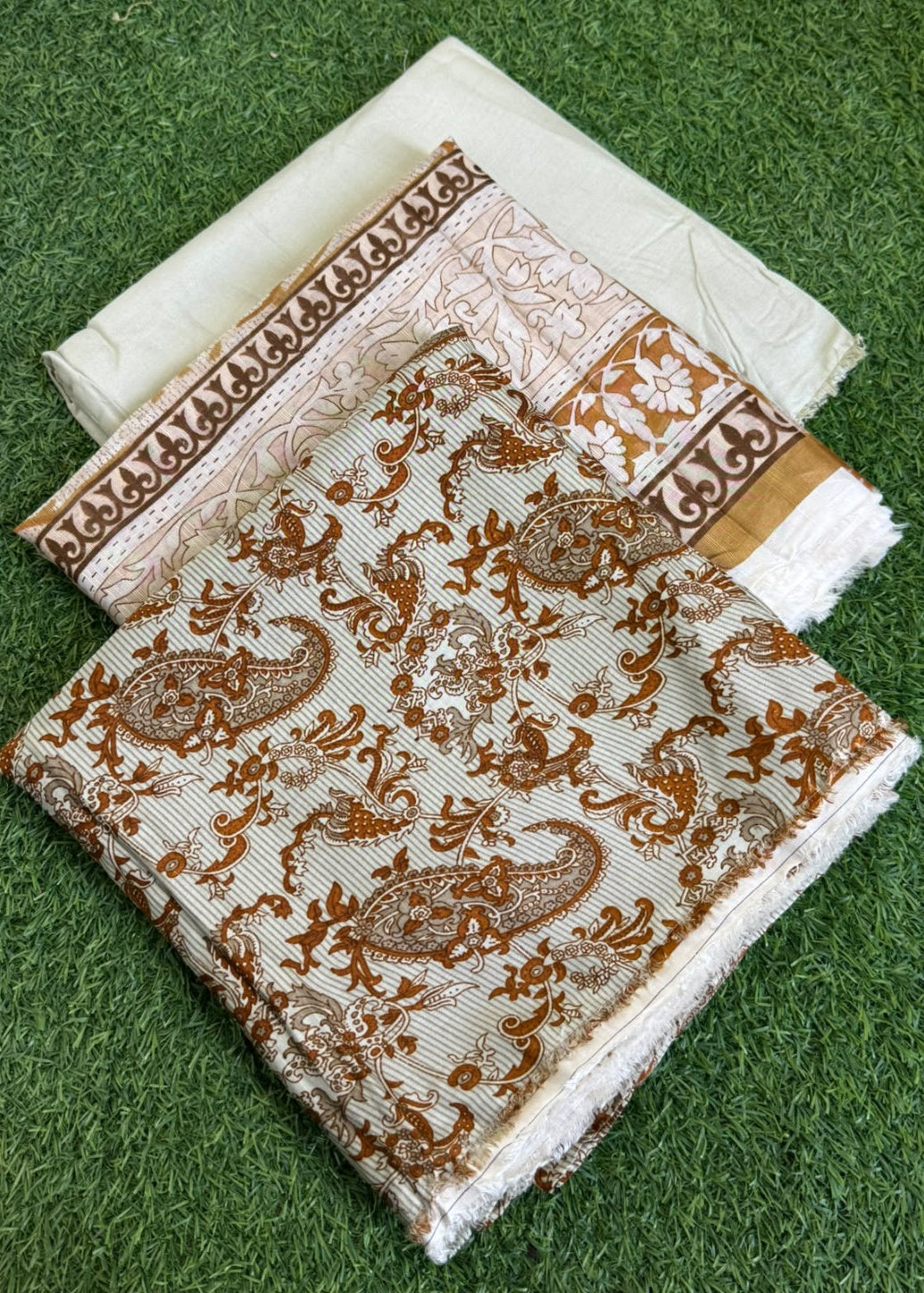 A folded set of traditional Indian fabric, likely cotton saree material, featuring a cream base with brown paisley and floral motifs, and a border print, displayed on artificial green grass.