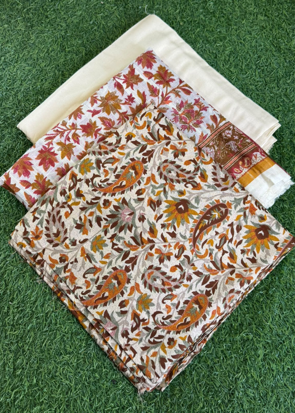 Three folded pieces of printed fabric laid on green grass. The front piece has a dense paisley and floral pattern in brown, orange, and beige tones, the middle piece has red and orange floral motifs on a white base, and the back piece is a solid cream color.