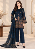 A woman models a navy blue three-piece salwar kameez suit with intricate gold embroidery, featuring a long-sleeve tunic, matching trousers, and a sheer dupatta, set against a warm beige background with decorative vases.