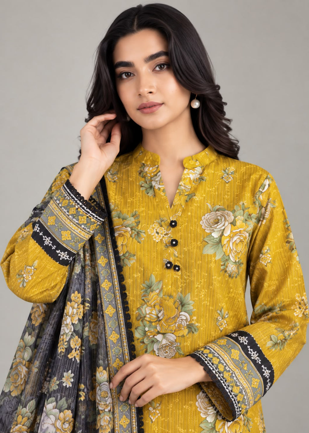 A woman modeling a mustard yellow floral kurta with black buttons and a matching dark gray dupatta featuring yellow and green floral patterns and black border detailing, set against a solid gray studio background.